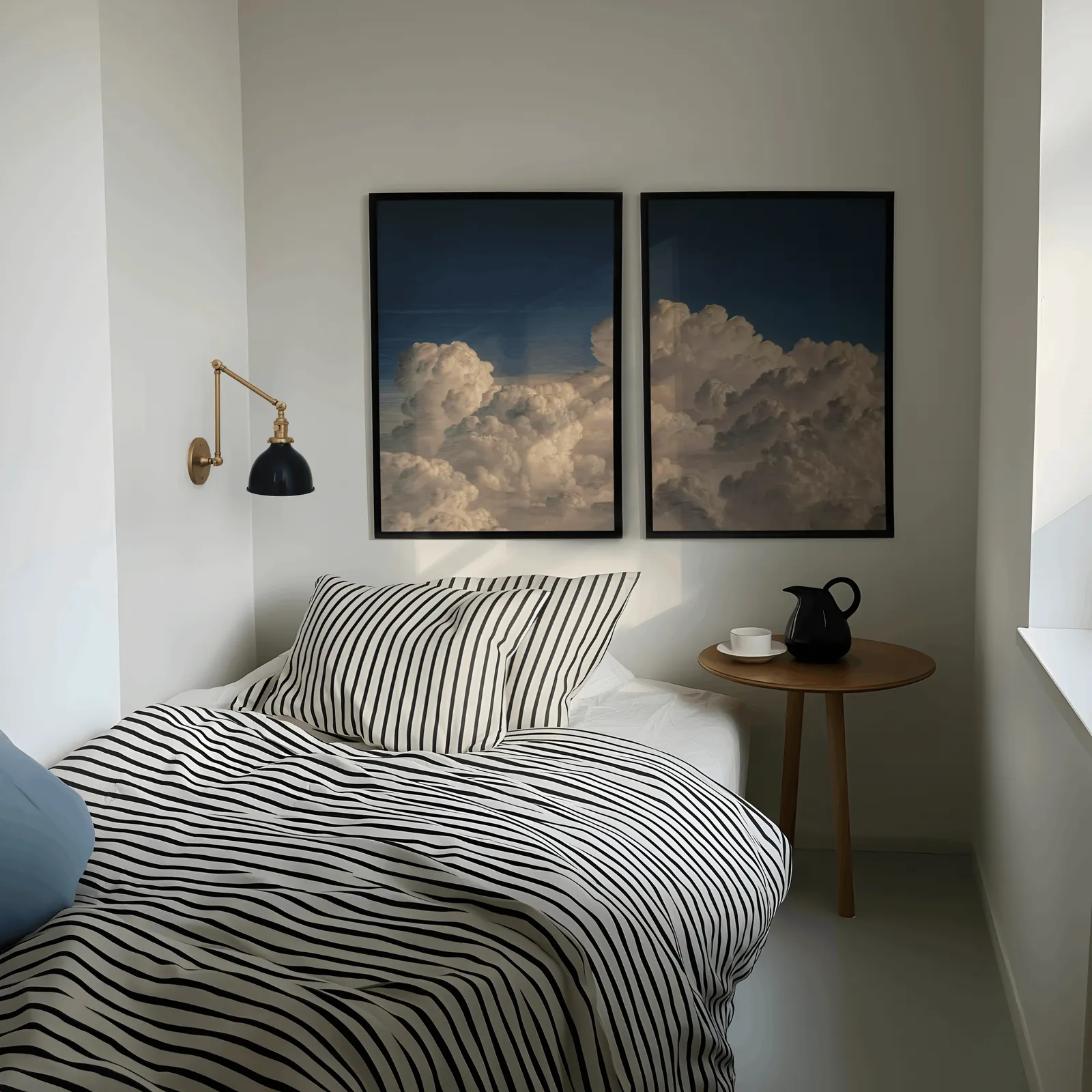 blue sky cloud landscape wall art set of two, above a striped bed beside a brass swing-arm lamp