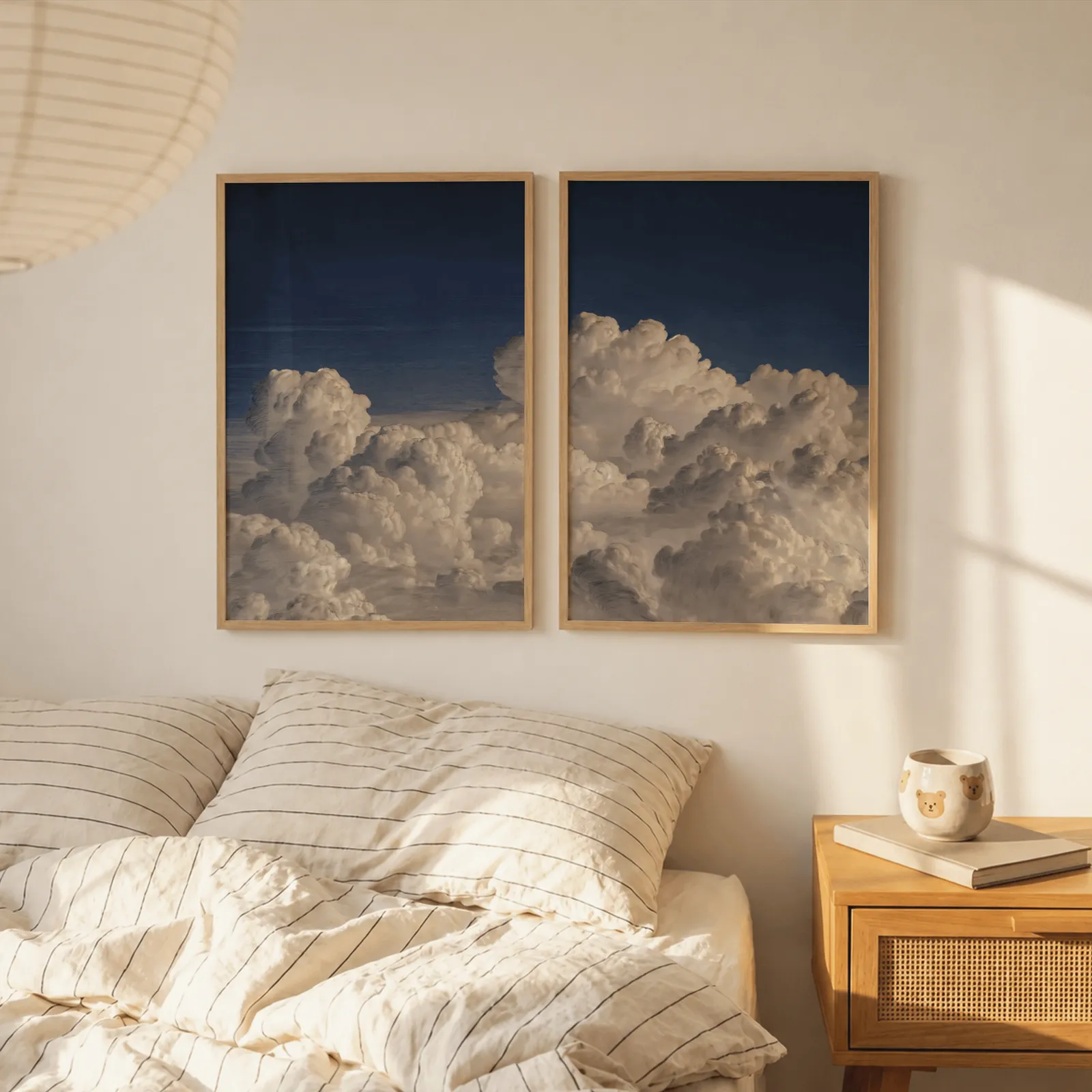 blue sky cloud landscape wall art set of two, above a striped linen bed beside a paper lantern shade