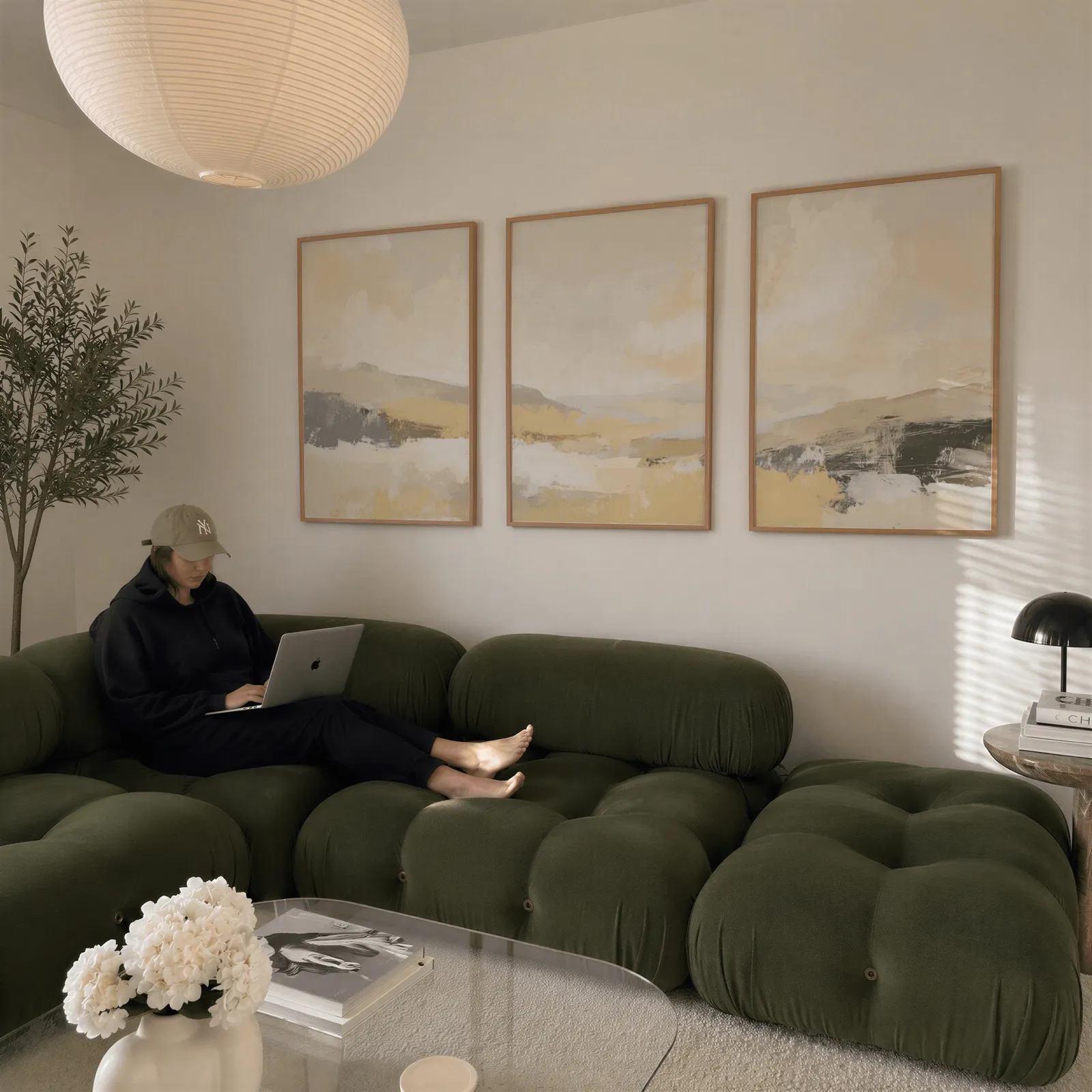beige abstract landscape triptych wall art, above a plush olive velvet sectional beneath a paper lantern light