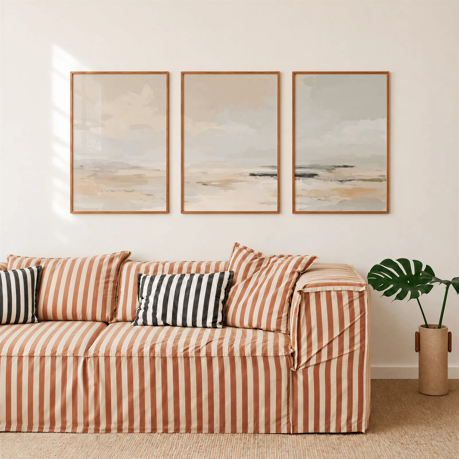 neutral coastal landscape triptych wall art, above a rust-striped sofa beside a potted monstera