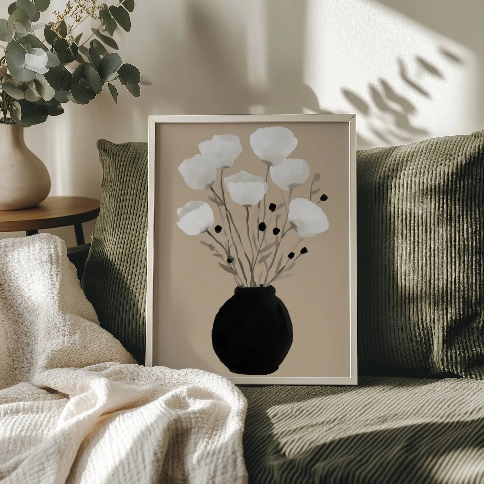 minimalist botanical wall art with black vase and white flowers, propped on an olive corduroy sofa beside a eucalyptus vase