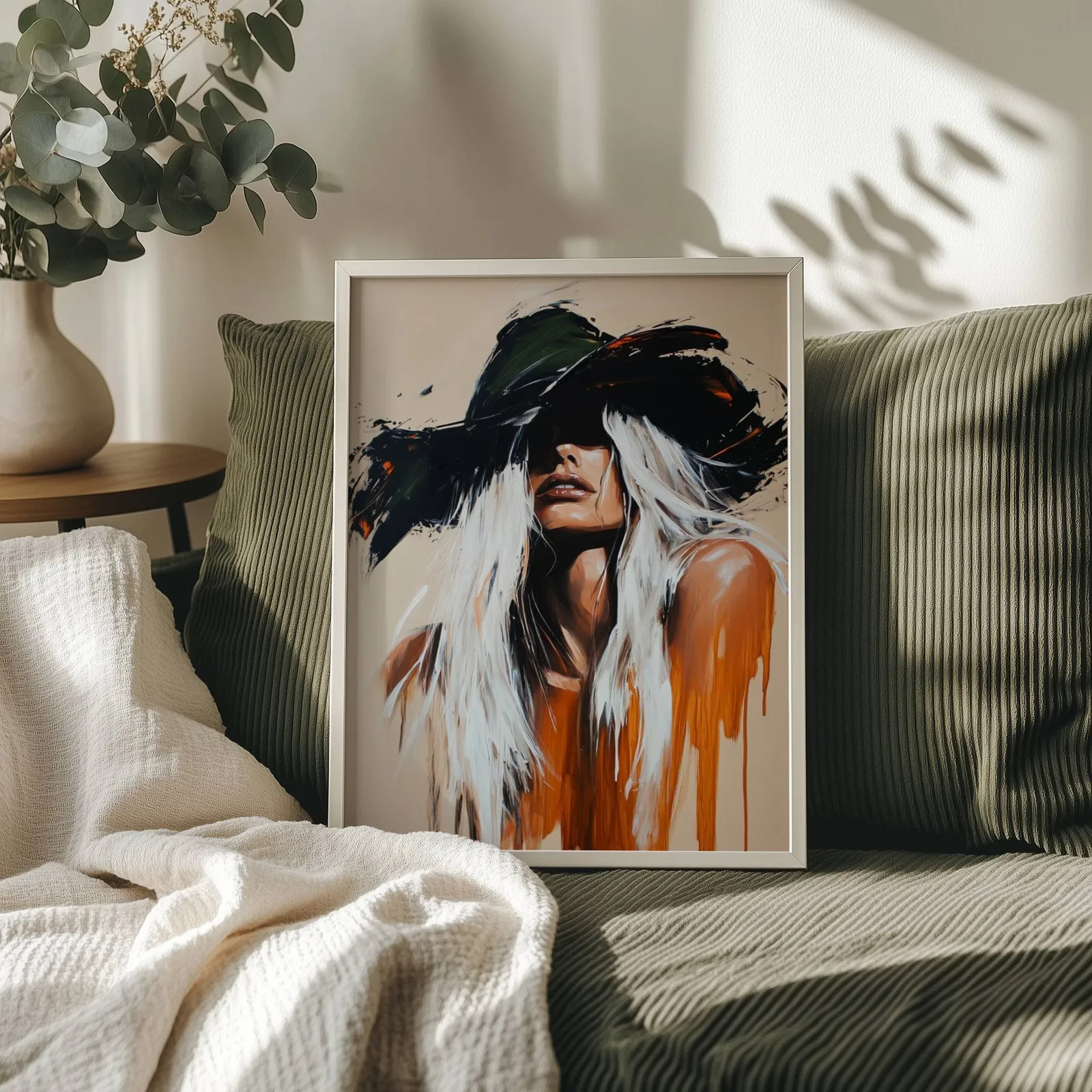 abstract cowgirl wall art in black and orange, propped on an olive corduroy sofa beside a eucalyptus vase