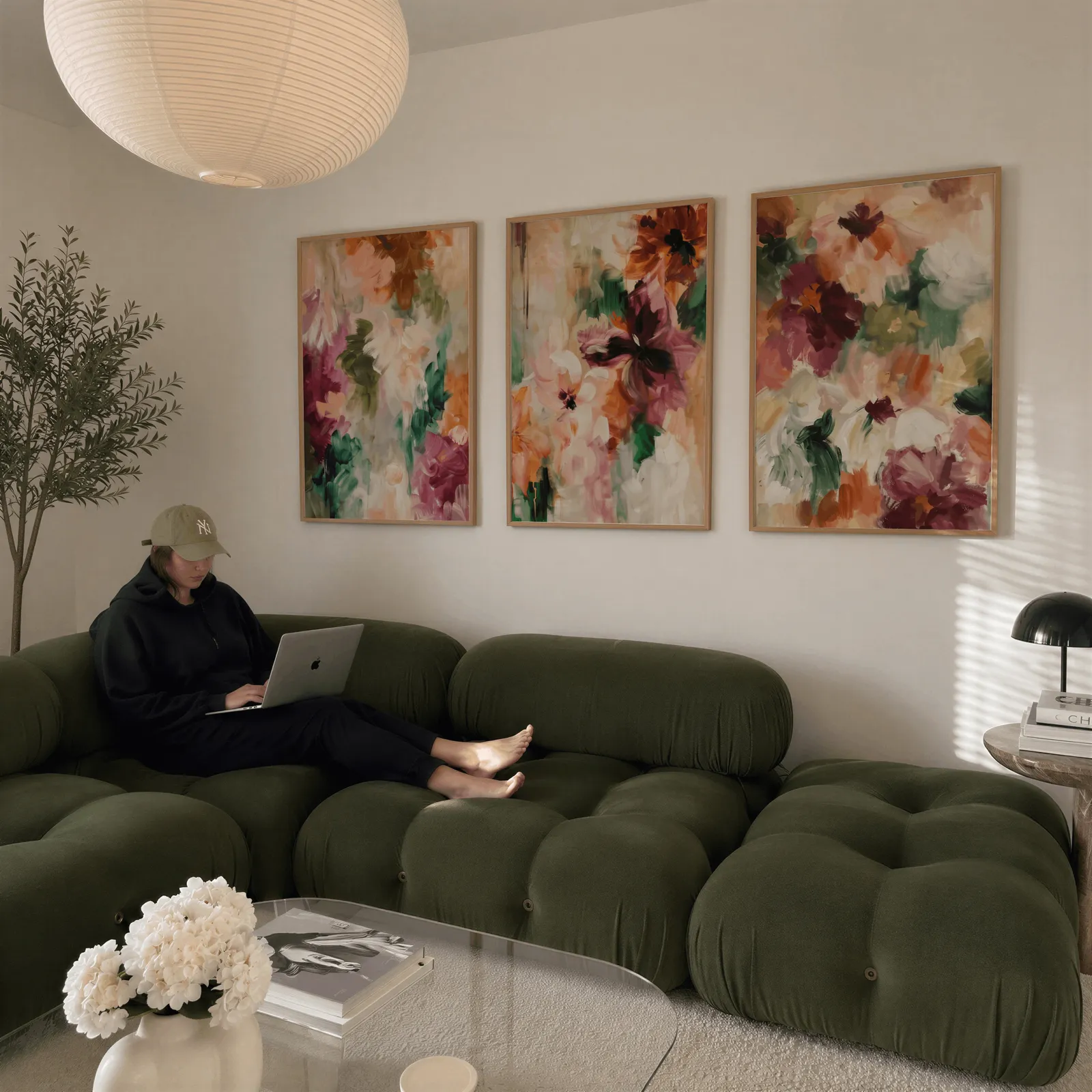 colorful floral triptych wall art in burgundy and green, above a plush olive velvet sectional beneath a paper lantern light