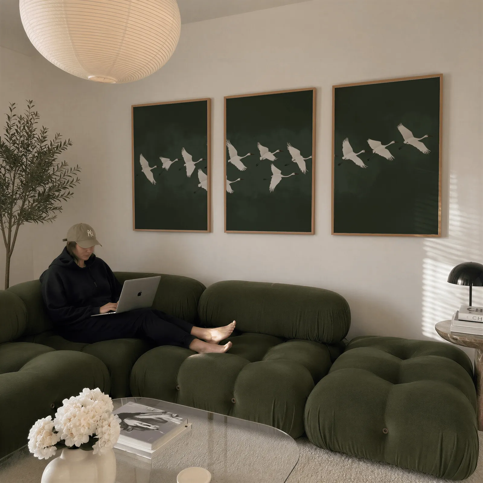 green crane wall art set of 3, above a plush olive velvet sectional beneath a paper lantern light
