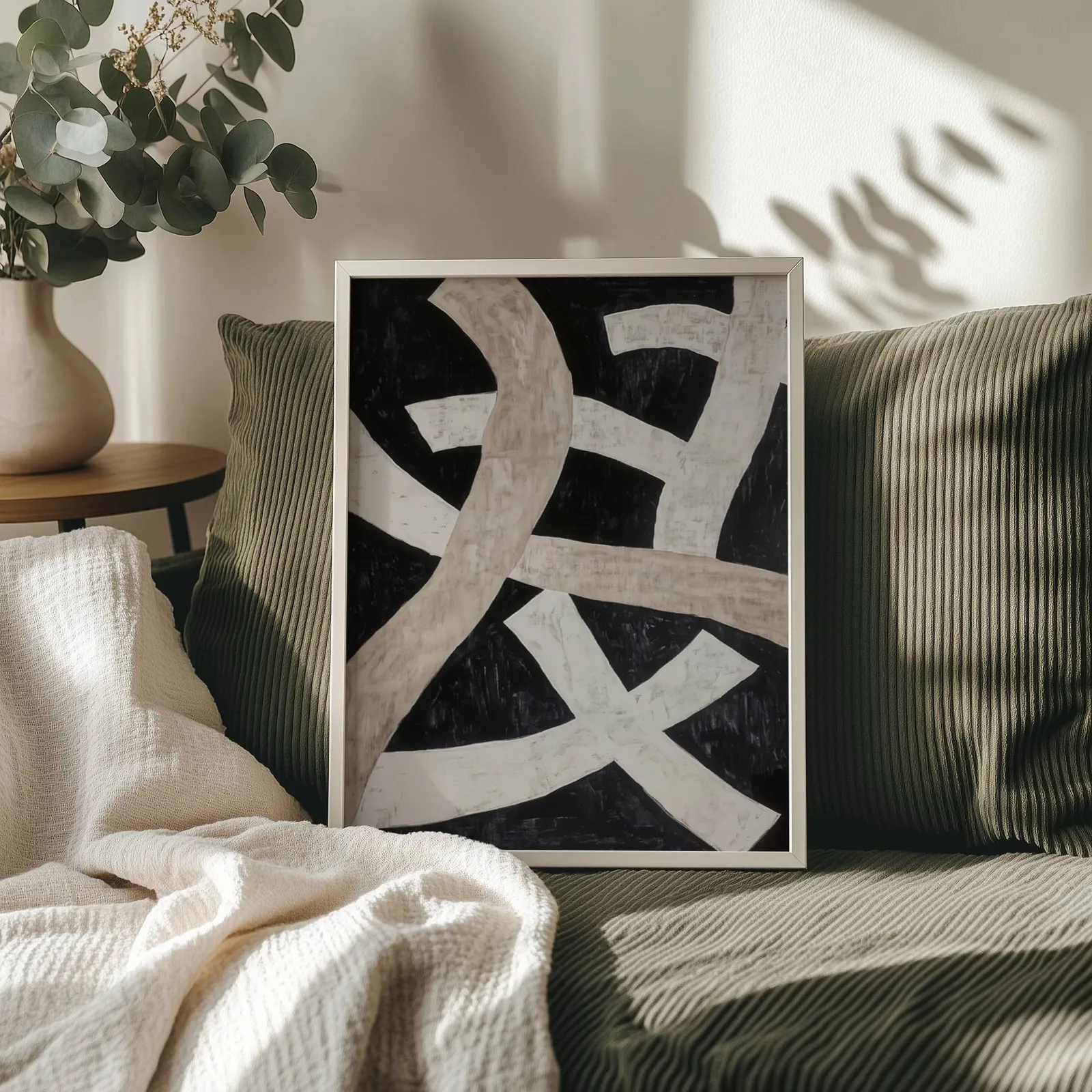 black minimalist abstract wall art with cream ribbons, propped on an olive corduroy sofa beside a eucalyptus vase