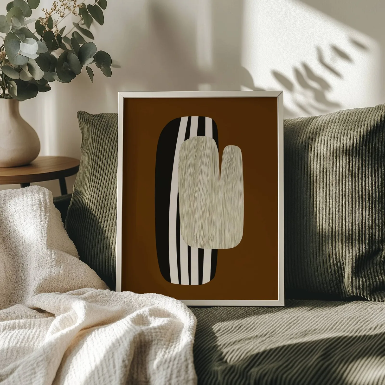 brown abstract wall art, propped on an olive corduroy sofa beside a eucalyptus vase