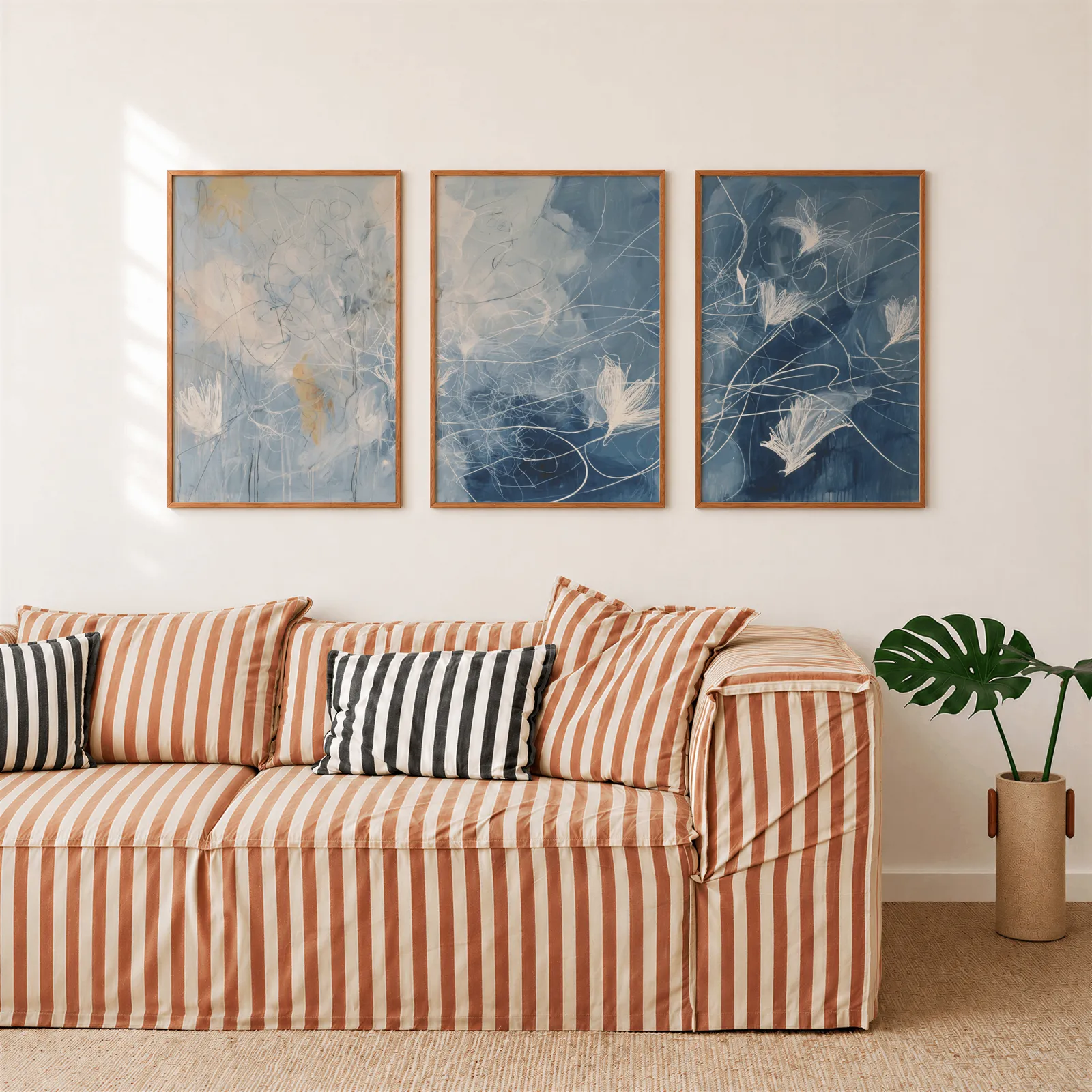 blue abstract floral triptych wall art, above a rust-striped sofa beside a potted monstera