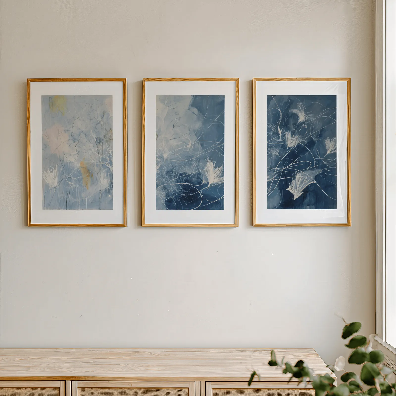 blue abstract floral triptych wall art, in a row above a light wooden sideboard near a window