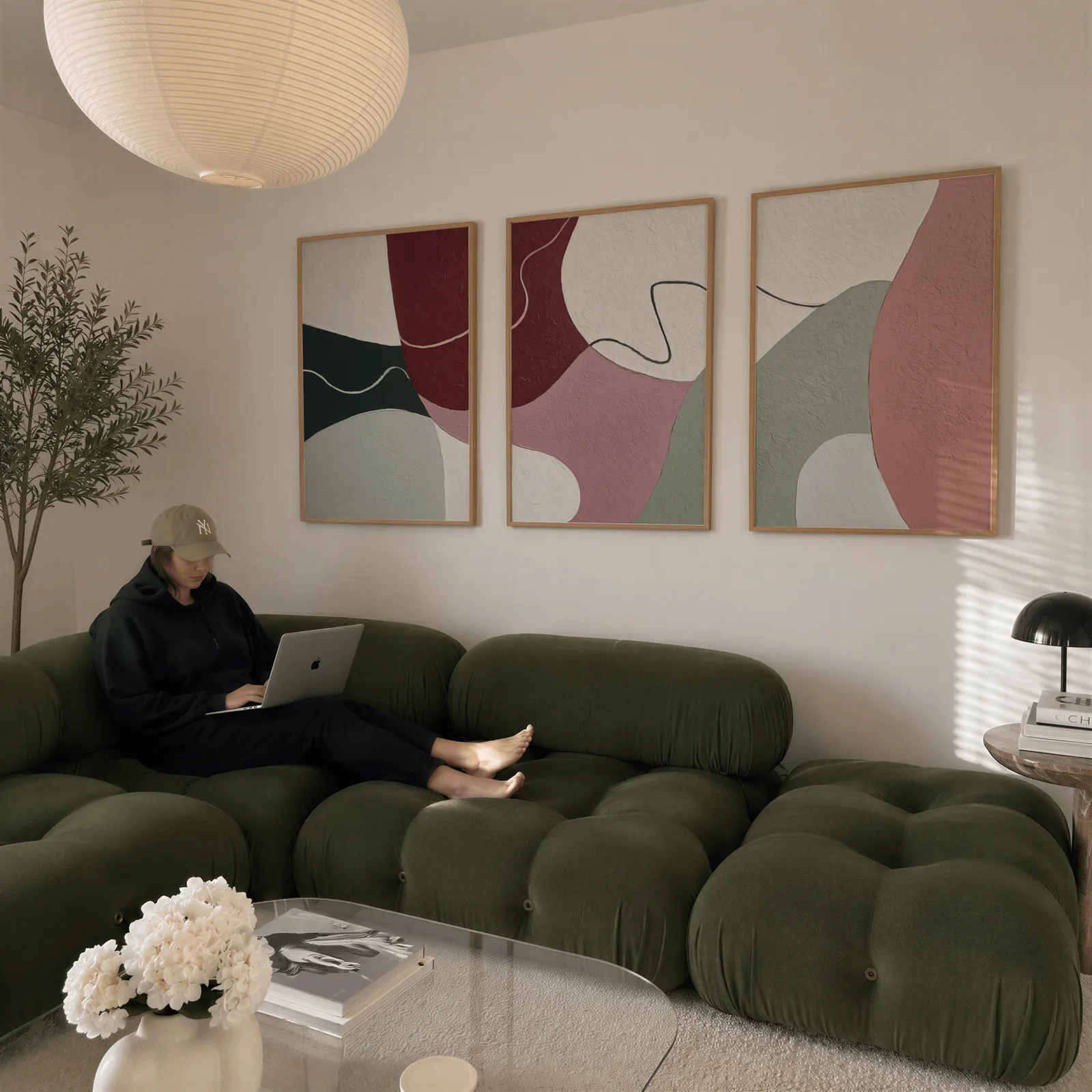 abstract triptych wall art in burgundy and pink, above a plush olive velvet sectional beneath a paper lantern light