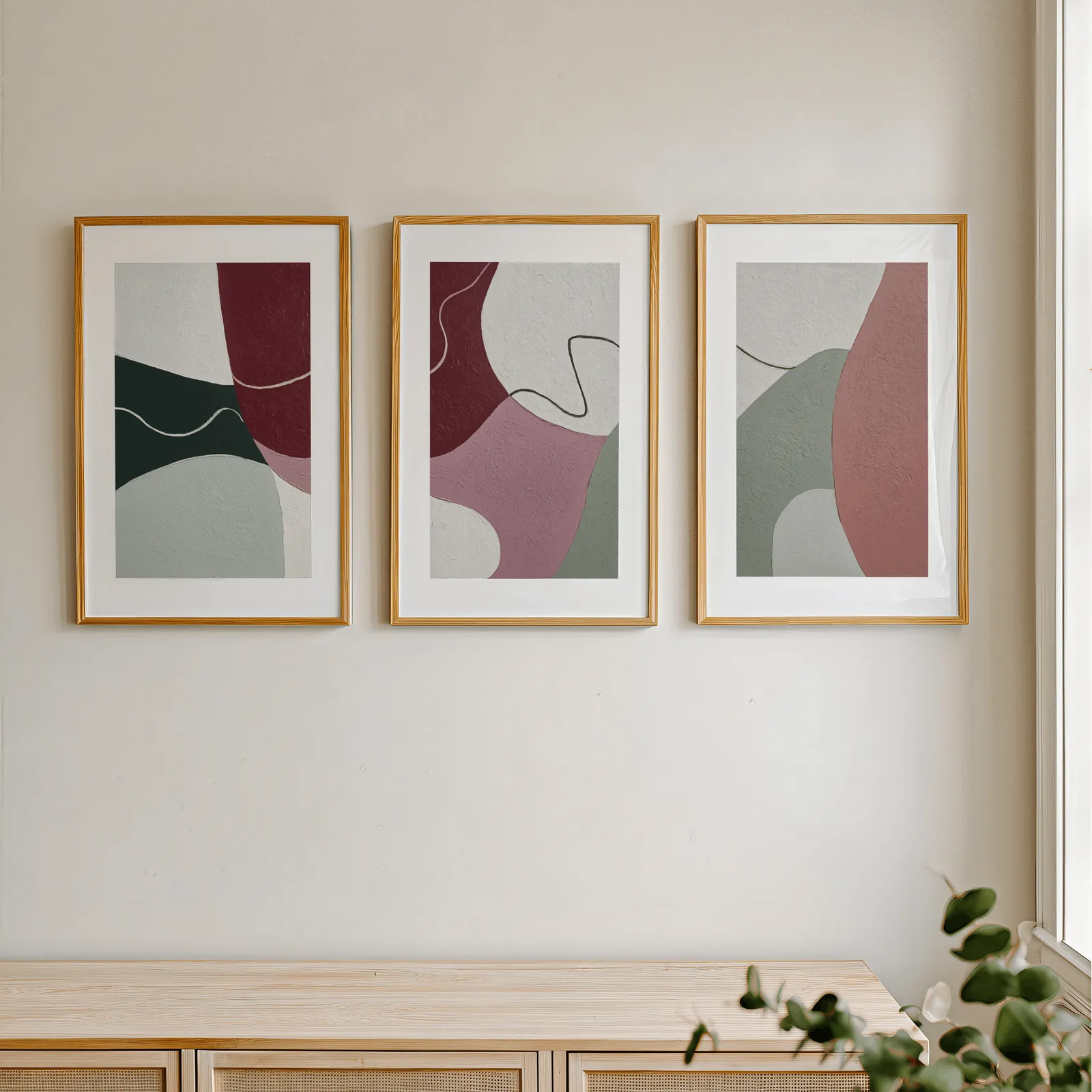 abstract triptych wall art in burgundy and pink, in a row above a light wooden sideboard near a window