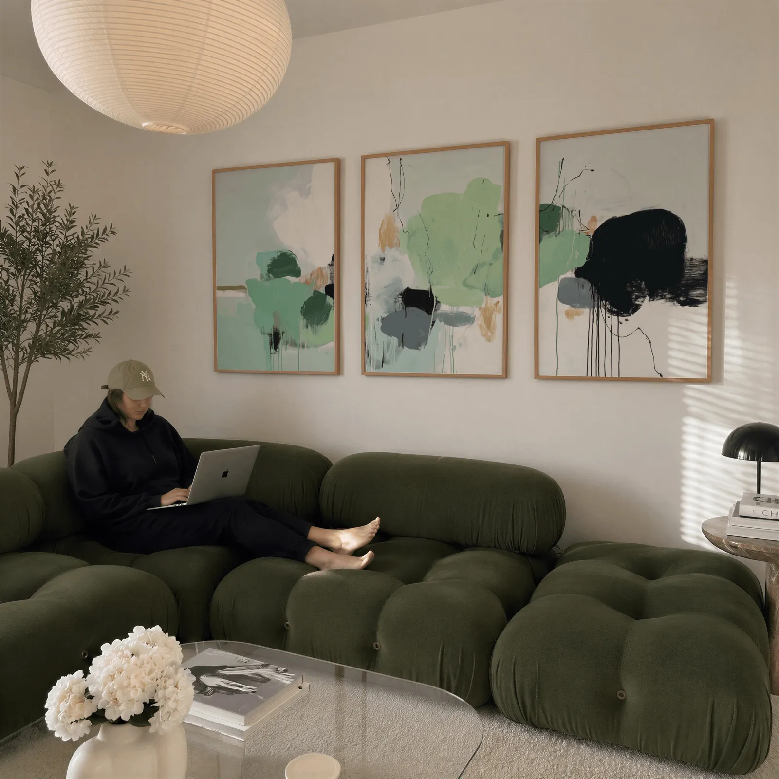 green abstract three piece wall art with black accents, above a plush olive velvet sectional beneath a paper lantern light