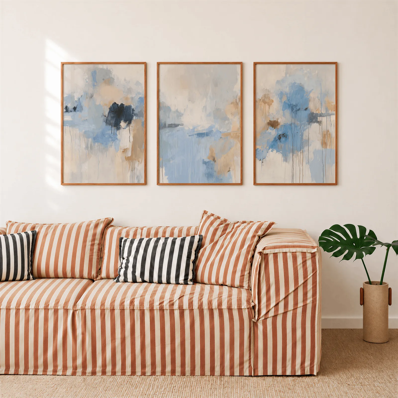 blue abstract three piece wall art, above a rust-striped sofa beside a potted monstera