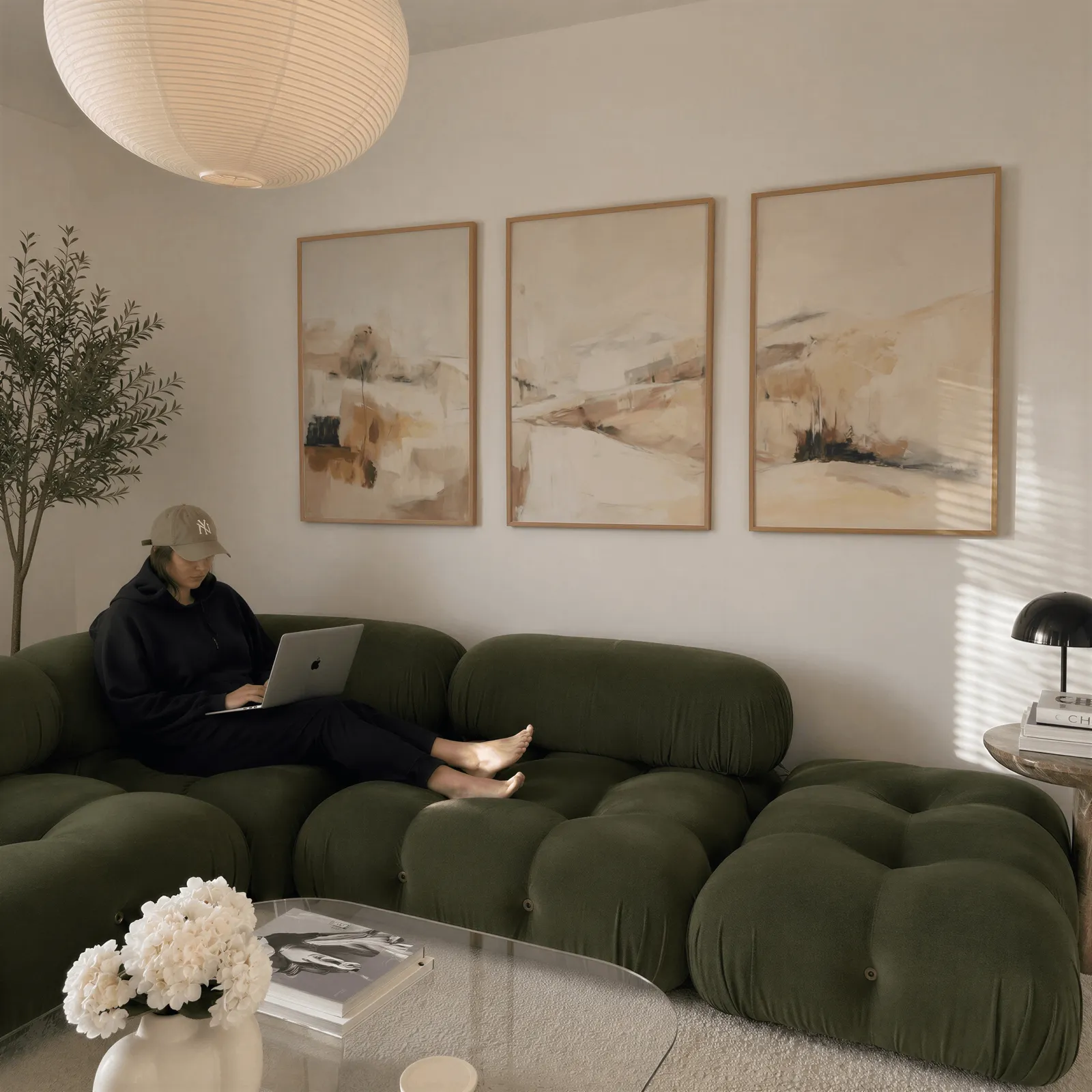 beige abstract landscape wall art set of three, above a plush olive velvet sectional beneath a paper lantern light