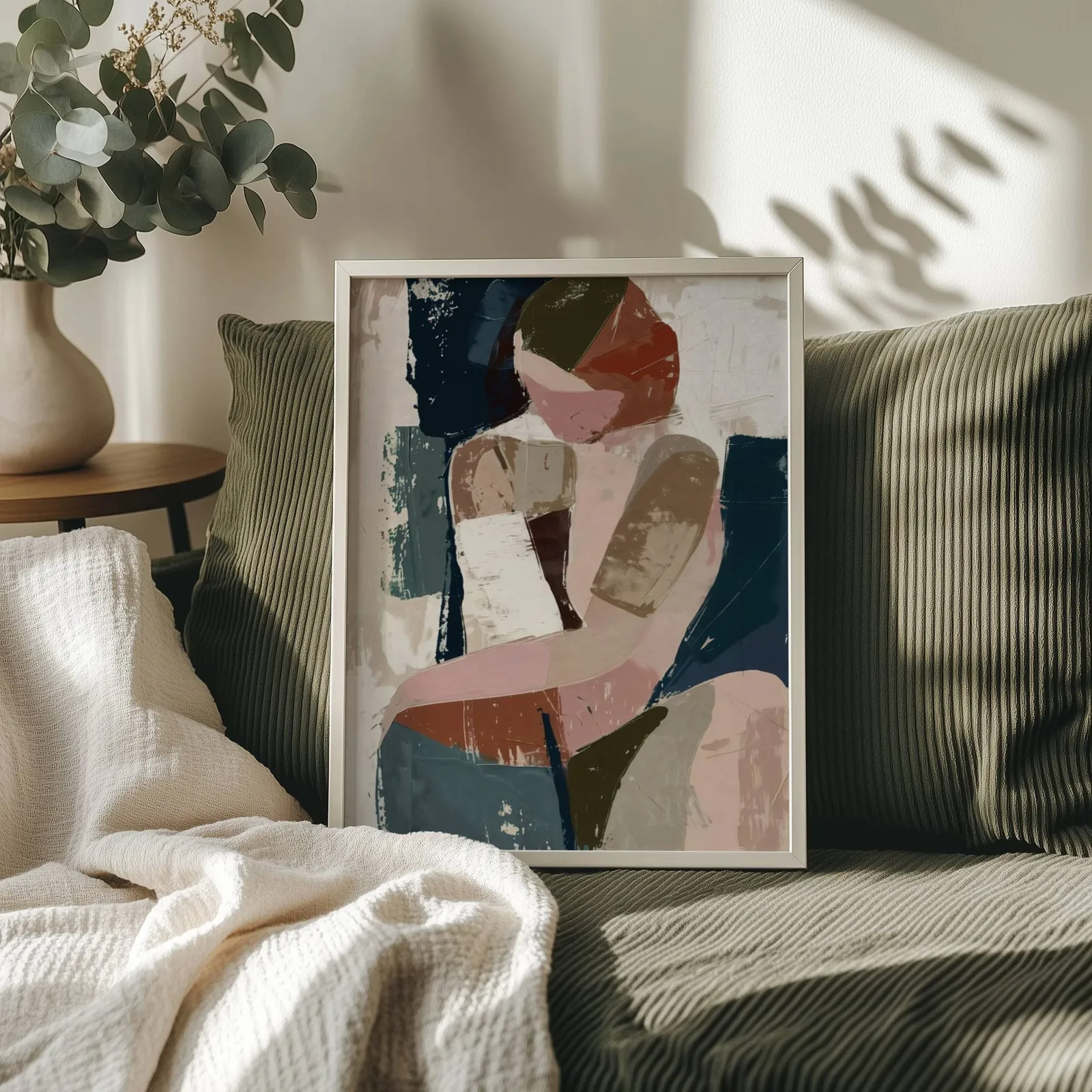 abstract face art in terracotta pink and navy, propped on an olive corduroy sofa beside a eucalyptus vase