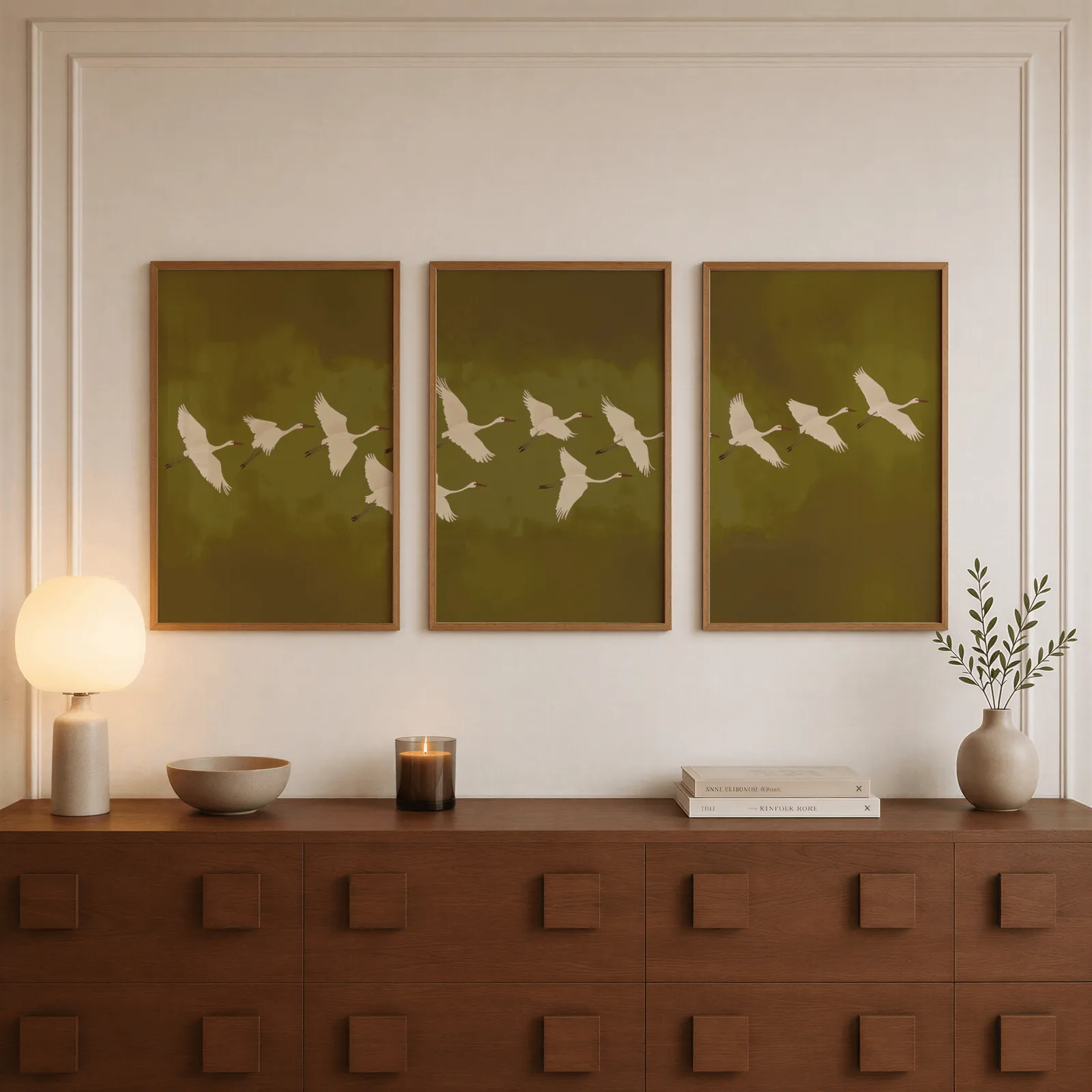 green cranes triptych wall art, above a walnut sideboard with a lit candle and glowing lamp