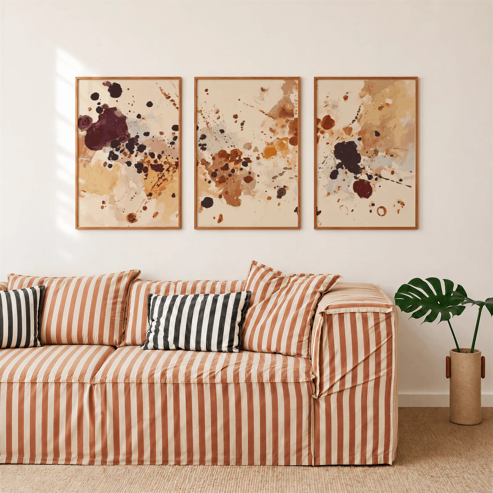 burgundy and brown abstract splatter wall art set of 3, above a rust-striped sofa beside a potted monstera