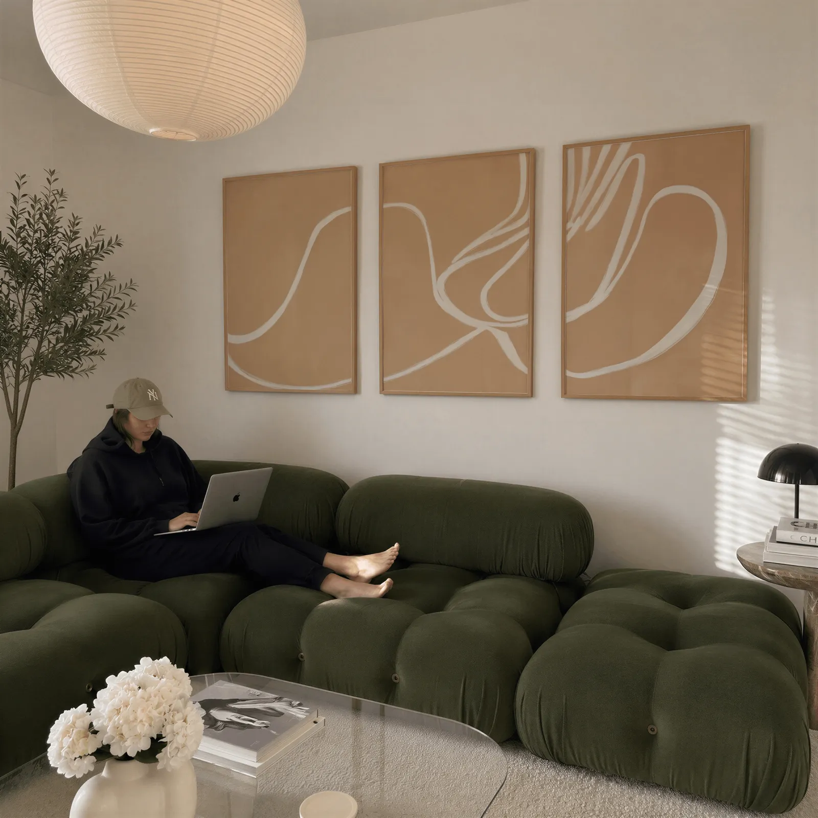beige abstract minimalist wall art set of three, above a plush olive velvet sectional beneath a paper lantern light