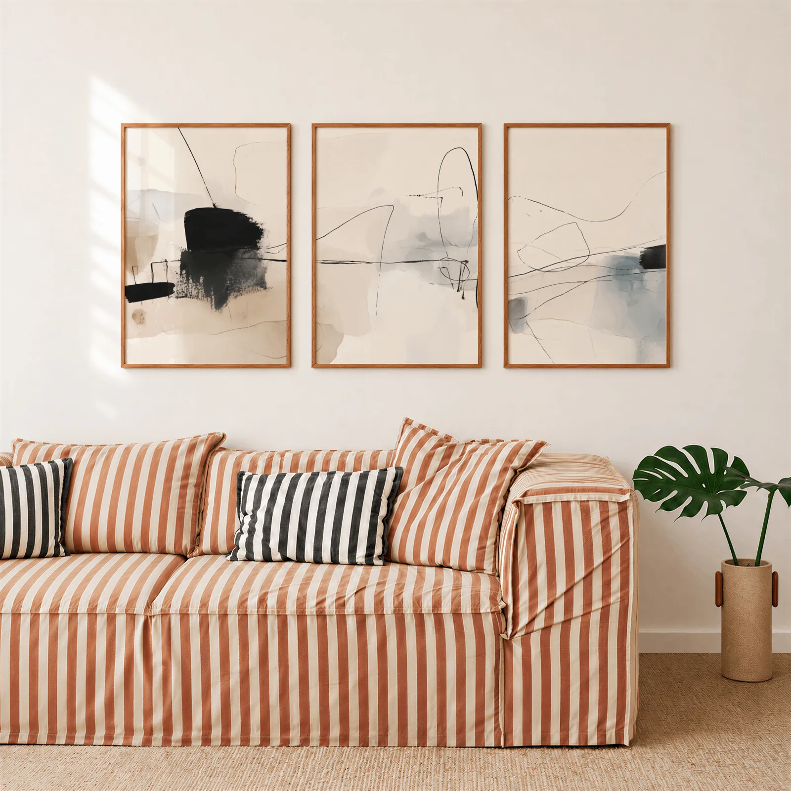 neutral abstract minimalist wall art set of three, above a rust-striped sofa beside a potted monstera