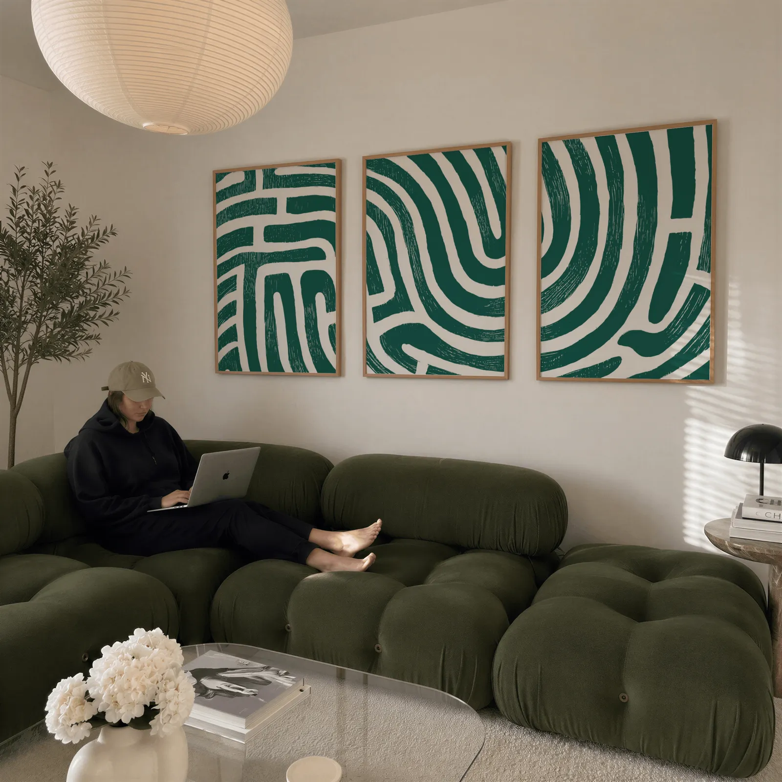 green abstract maze line triptych wall art, above a plush olive velvet sectional beneath a paper lantern light