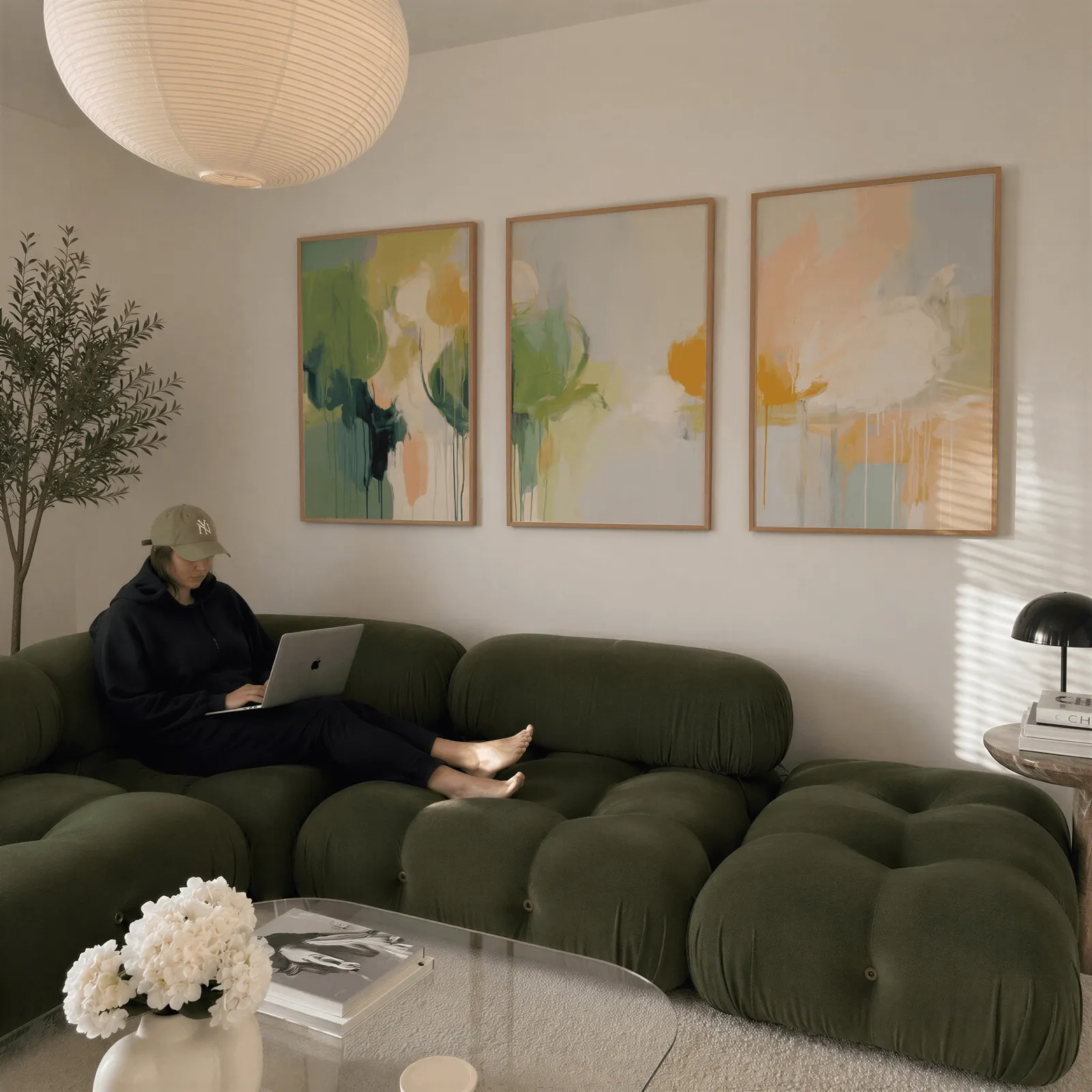green and orange abstract nature triptych wall art, above a plush olive velvet sectional beneath a paper lantern light