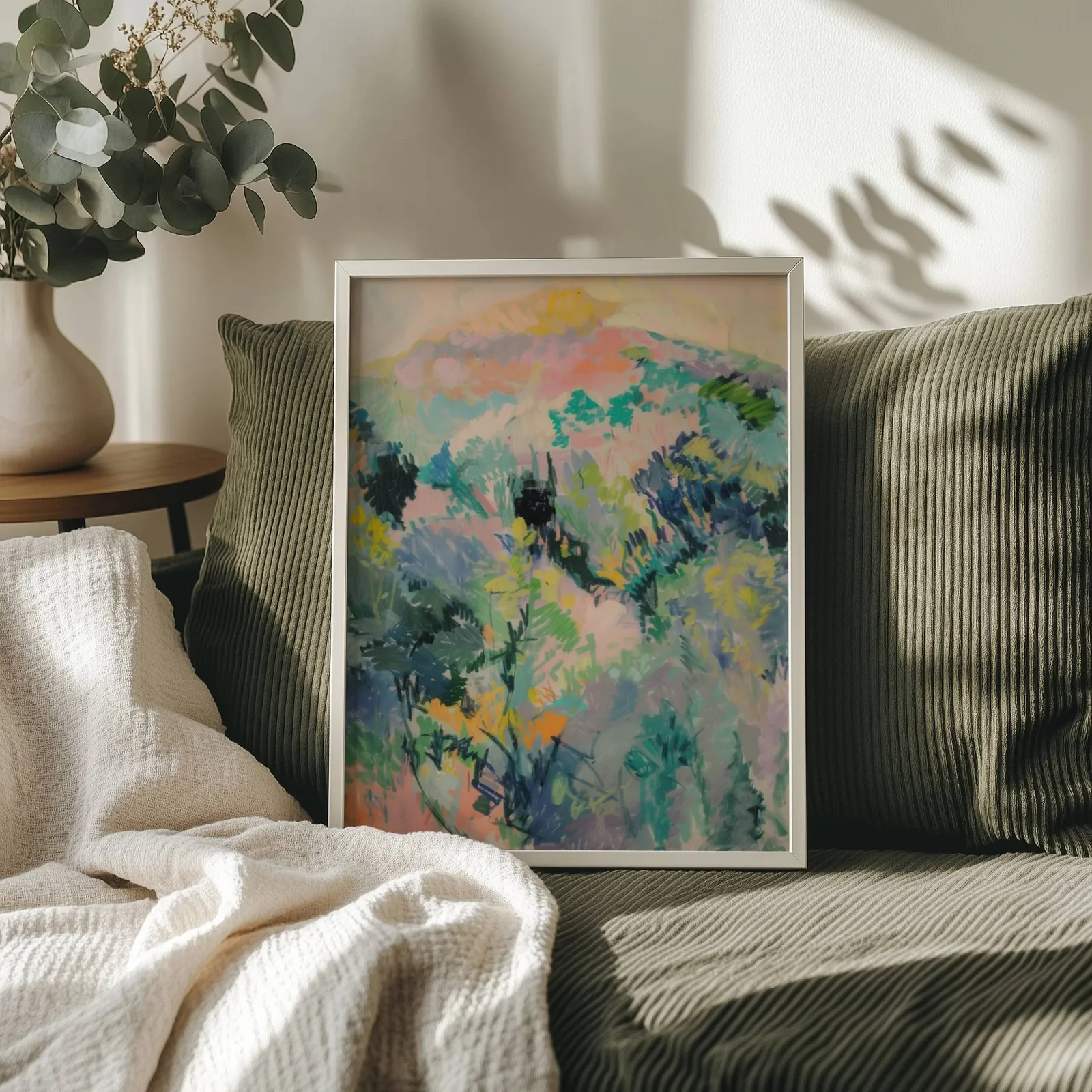colorful landscape wall art of hillside foliage, propped on an olive corduroy sofa beside a eucalyptus vase