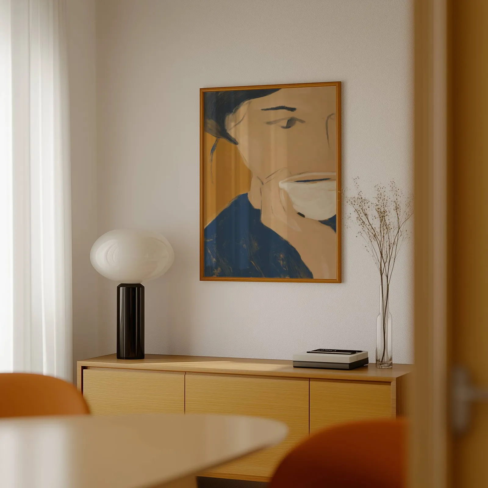 abstract portrait wall art with teacup in blue and orange, above a light wood sideboard beside a bulbous black lamp