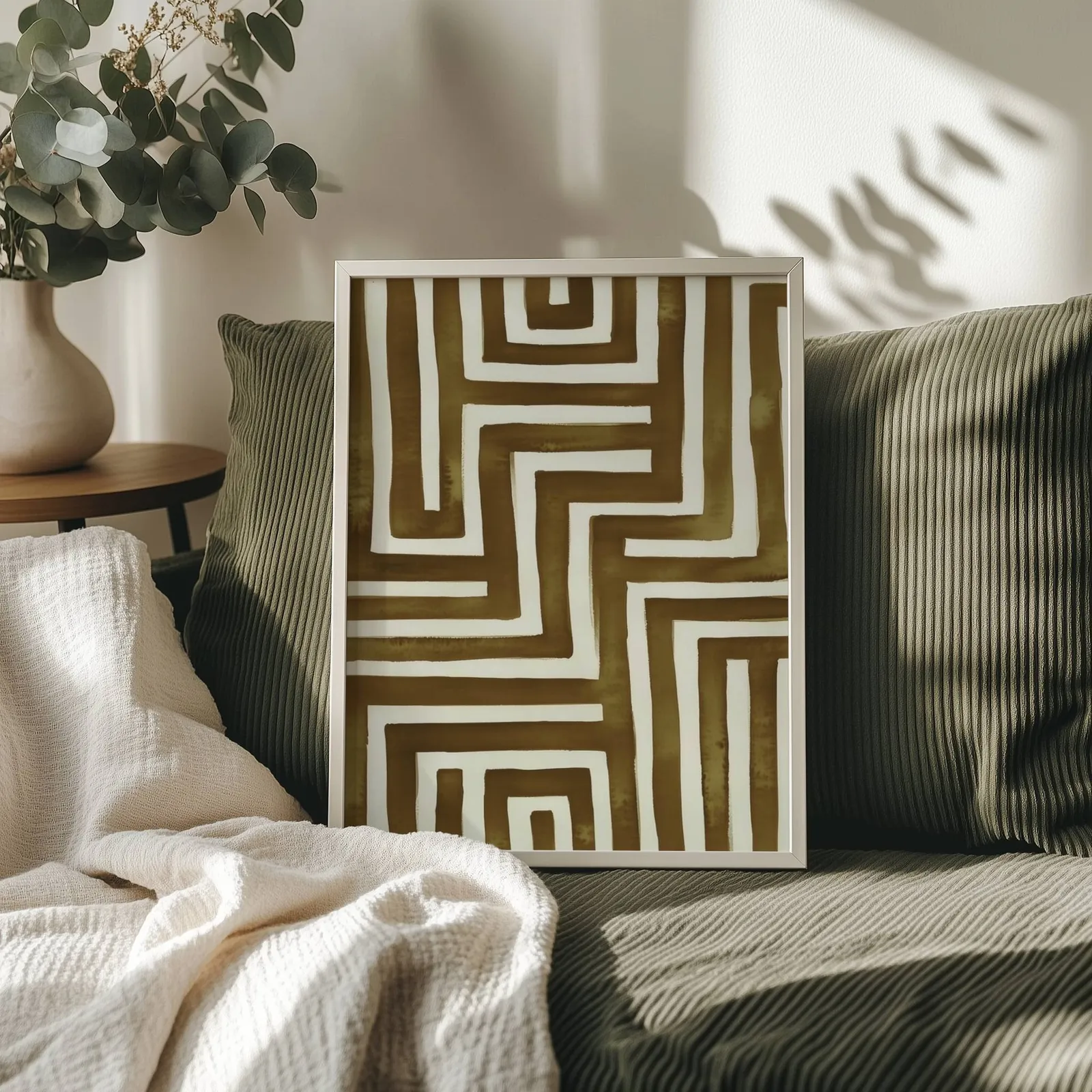 brown abstract maze wall art, propped on an olive corduroy sofa beside a eucalyptus vase