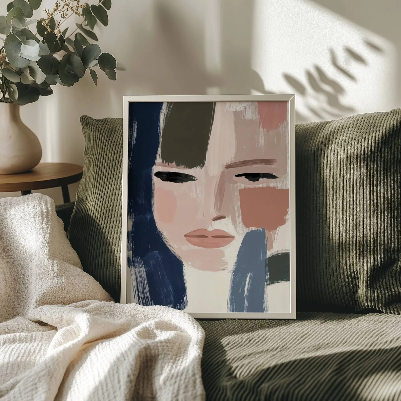 abstract woman face art in blue and terracotta, propped on an olive corduroy sofa beside a eucalyptus vase