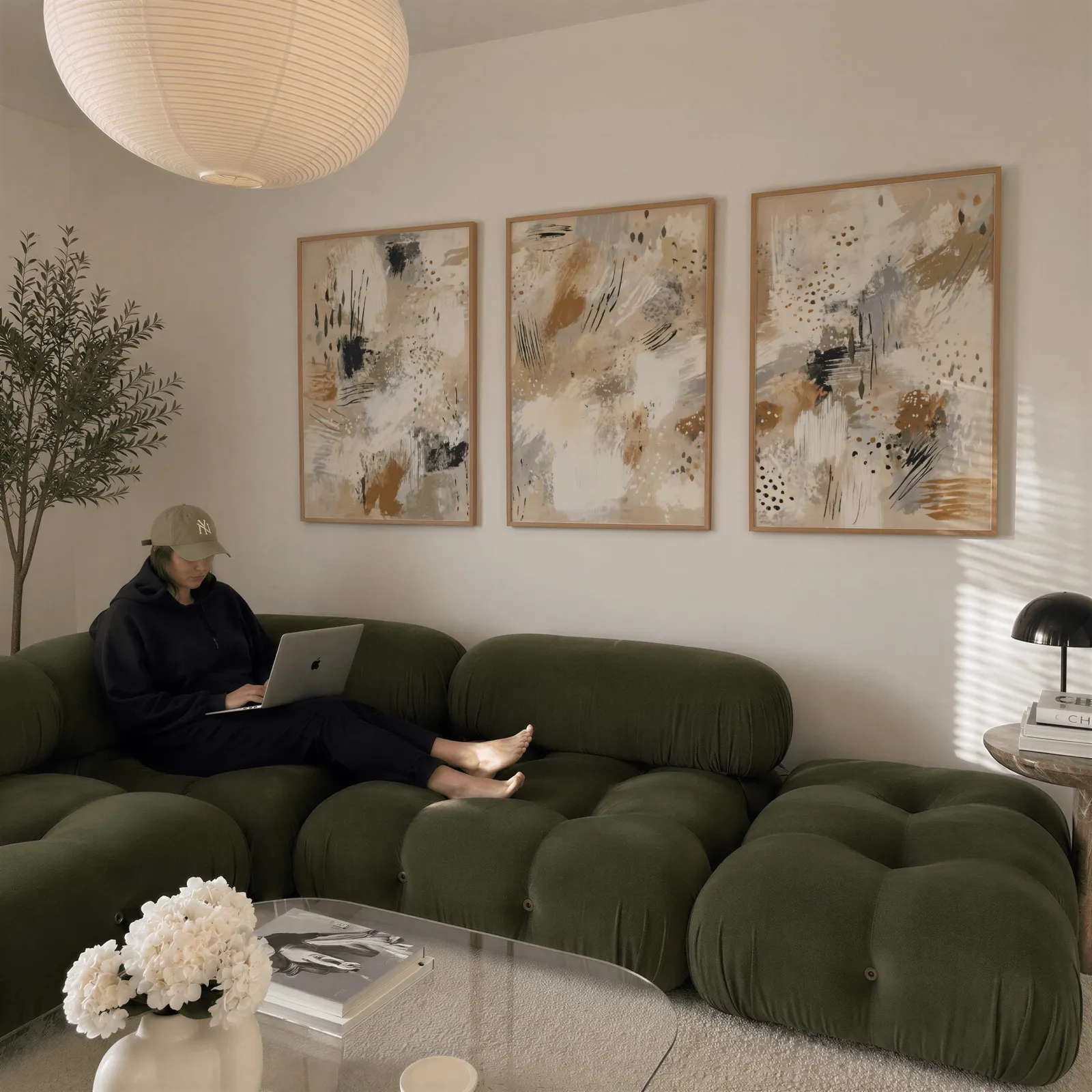 neutral abstract three piece wall art in beige and black, above a plush olive velvet sectional beneath a paper lantern light