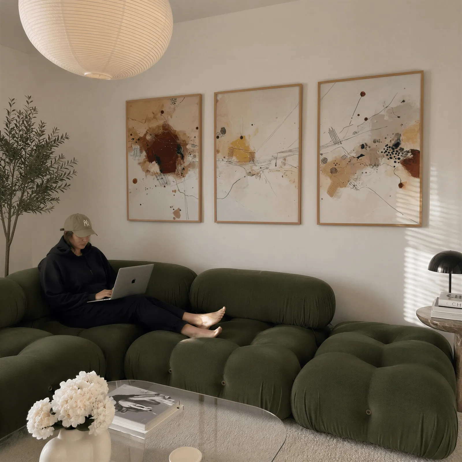 neutral abstract three piece wall art in brown and beige, above a plush olive velvet sectional beneath a paper lantern light