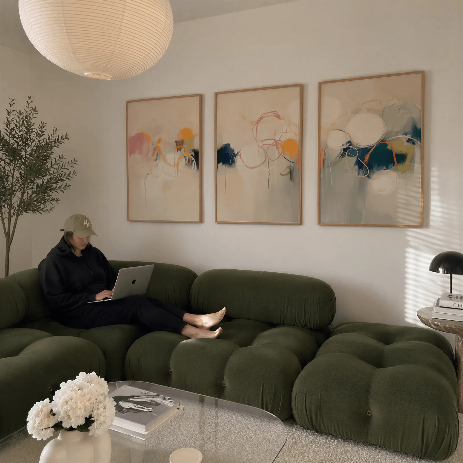colorful abstract triptych wall art in pink orange and blue, above a plush olive velvet sectional beneath a paper lantern light