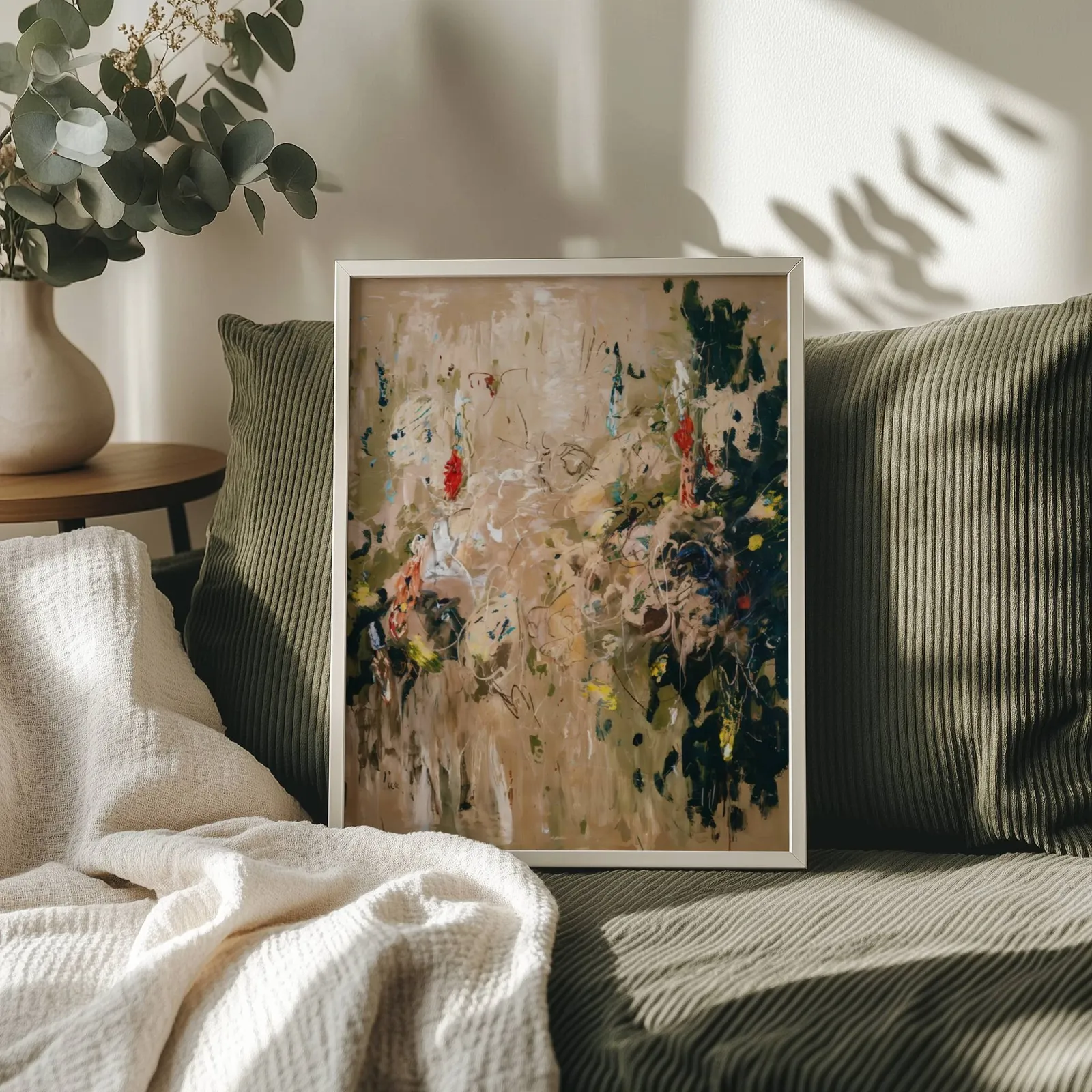 abstract floral wall art in beige and green, propped on an olive corduroy sofa beside a eucalyptus vase