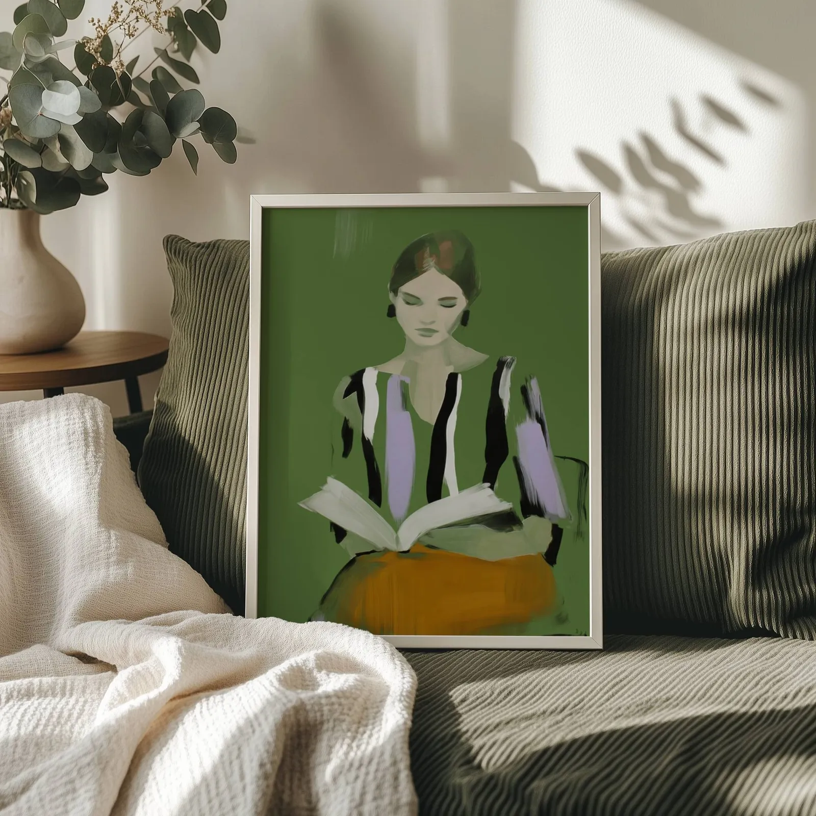 green modern woman reading wall art, propped on an olive corduroy sofa beside a eucalyptus vase